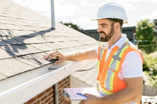 Roofing contractor inspecting asphalt shingles and gutter while documenting roof condition for warranty assessment